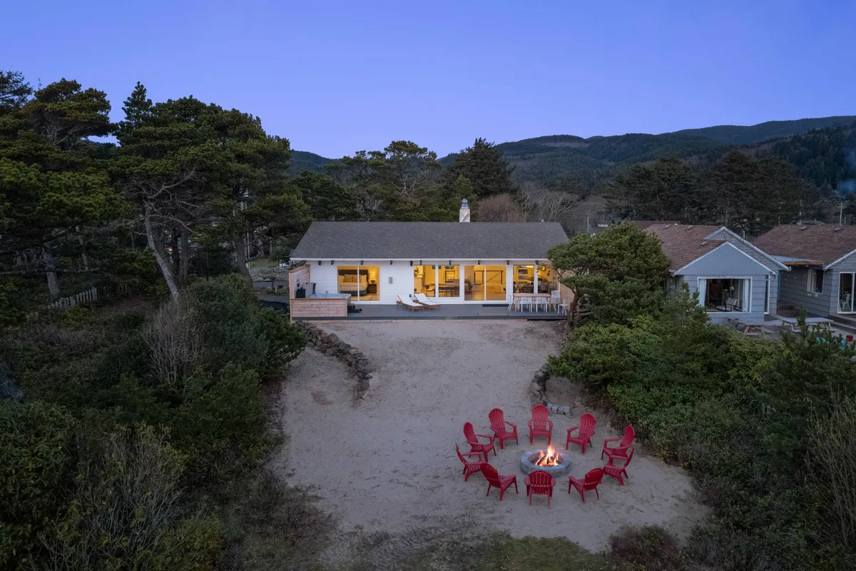 An inviting exterior shot featuring the house, a cozy fire pit surrounded by red chairs, and a backdrop of towering trees. The scene suggests warm social gatherings, storytelling, and enjoying the outdoors at dusk or night.