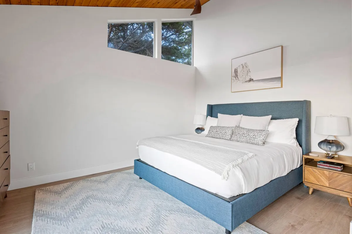 A serene bedroom space featuring a large bed with soft bedding, flanked by nightstands and illuminated by gentle natural light streaming through adjacent windows. Artwork above the bed adds a touch of character, and the calm setting is ideal for rest and quiet moments.