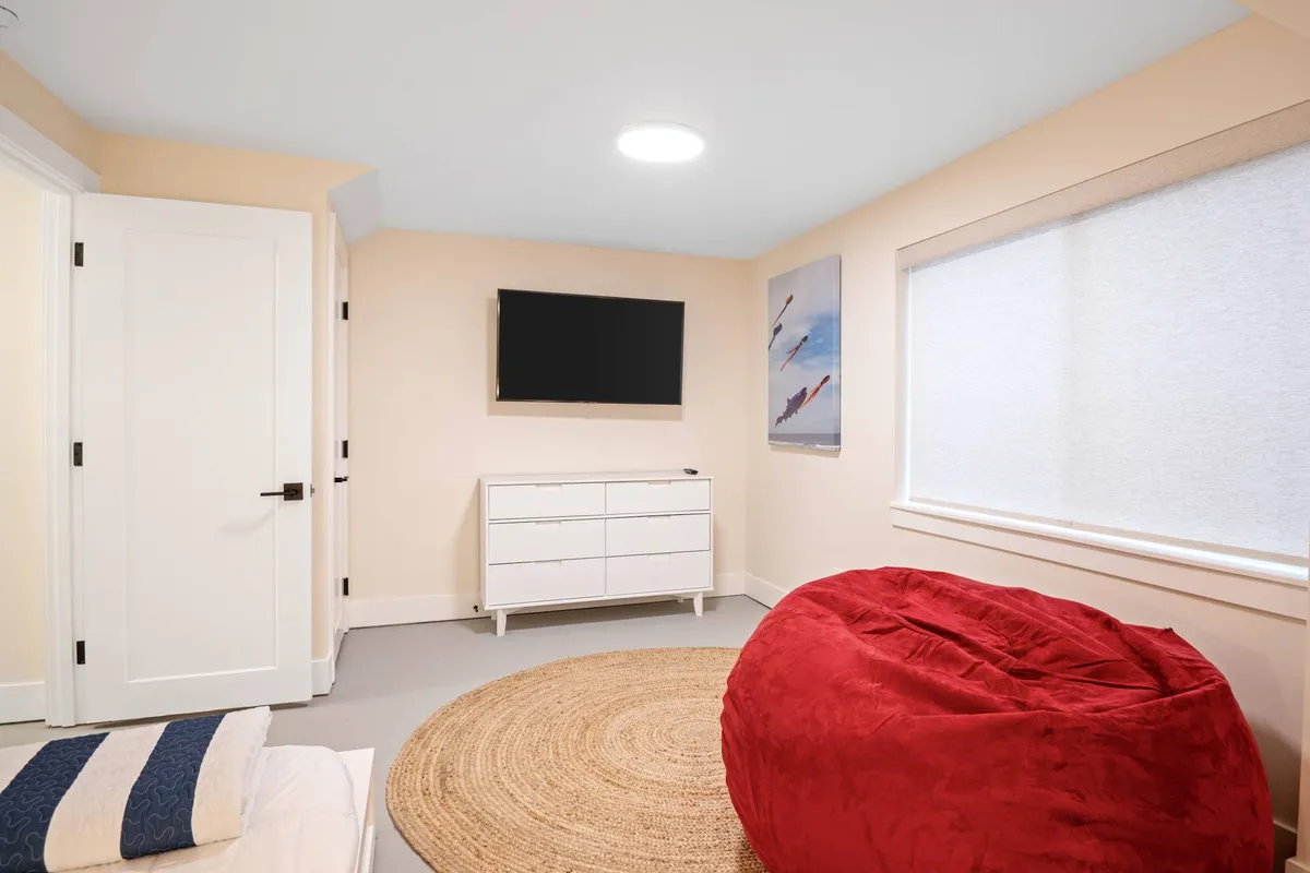 A cozy lower-level family room featuring a vibrant red bean bag chair, a white dresser, and a large flat-screen TV mounted on the wall. Artwork adorns the walls, and plush seating creates an inviting space for relaxation, watching TV, or playing games. The atmosphere is casual and kid-friendly, ideal for unwinding after a day of adventure.
