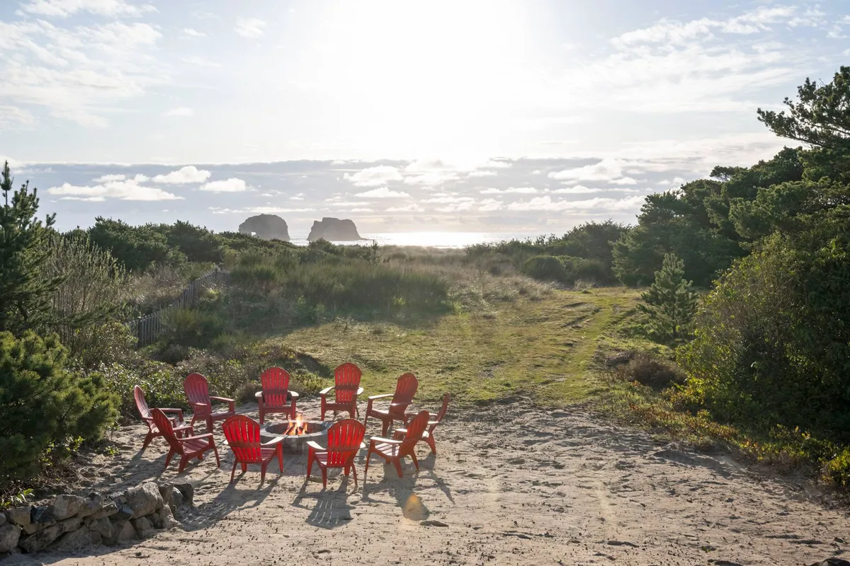 A close view of a fire pit surrounded by classic red Adirondack chairs, with a stunning ocean view and rocky outcrops in the background. The scene is ideal for gatherings, sunset watching, and cozy evenings around the fire.