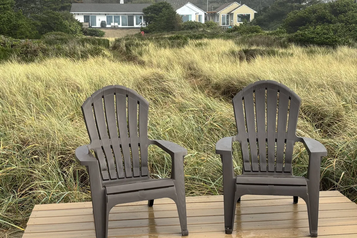Two Adirondack chairs positioned in dune grass overlook a tranquil stretch of beach, with two beach houses visible in the background. The scene exudes peacefulness and an invitation to relax in the open, natural landscape.