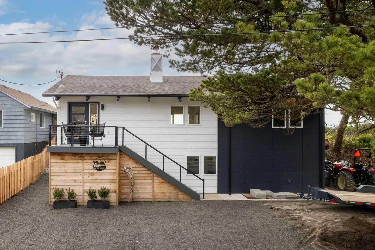 This image features the modern front design of the house, a porch with comfortable seating, and steps leading up to the entrance, surrounded by textured wood paneling. The setting offers an immediate sense of arrival and connection to the outdoors, inviting guests to pause and soak in the coastal vibe.