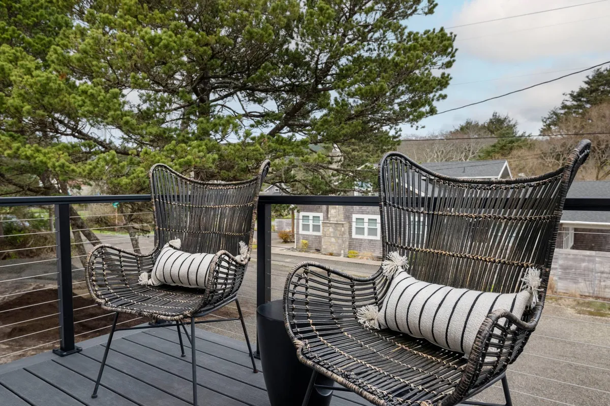 A peaceful balcony scene with two wicker chairs, decorative pillows, and surrounding trees. The morning light filters through the branches, suggesting the ideal spot for sipping coffee and taking in the ocean air or sunset views.