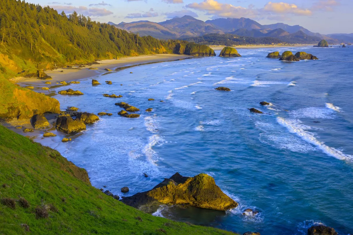 A dramatic coastal scene showcasing waves crashing on the beach, iconic rock formations, distant mountains, and forested cliffs. The image evokes a sense of exploration, natural beauty, and the allure of the Oregon Coast.