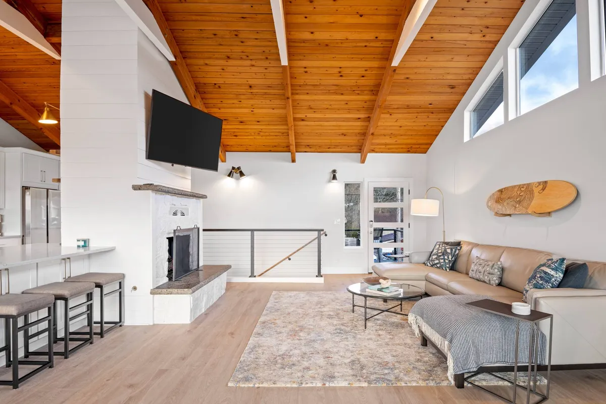 An inviting living room featuring a comfortable sofa, a cozy fireplace, wooden ceiling beams, a plush rug, and a television. Sunlight streams through windows, making it a welcoming spot for morning coffee, game nights, or movie marathons.