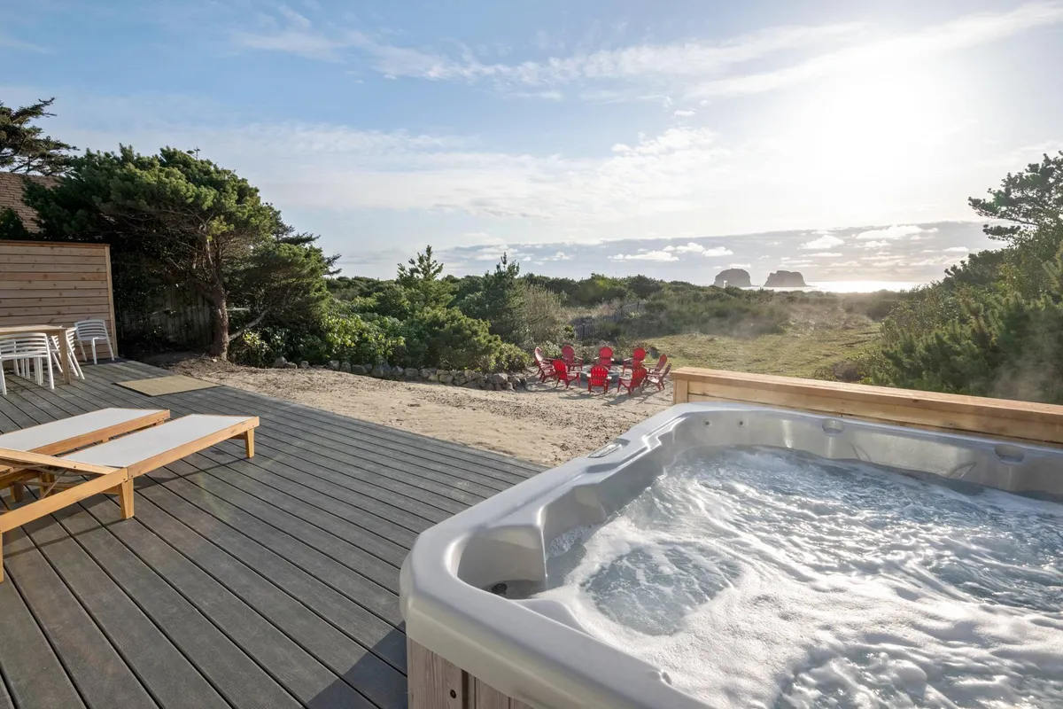 The hero image showcases an expansive deck overlooking rolling beach grass and the Pacific Ocean, featuring a steaming hot tub, inviting deck chairs, and a cozy fire pit—perfectly positioned for sunset views and social gatherings.