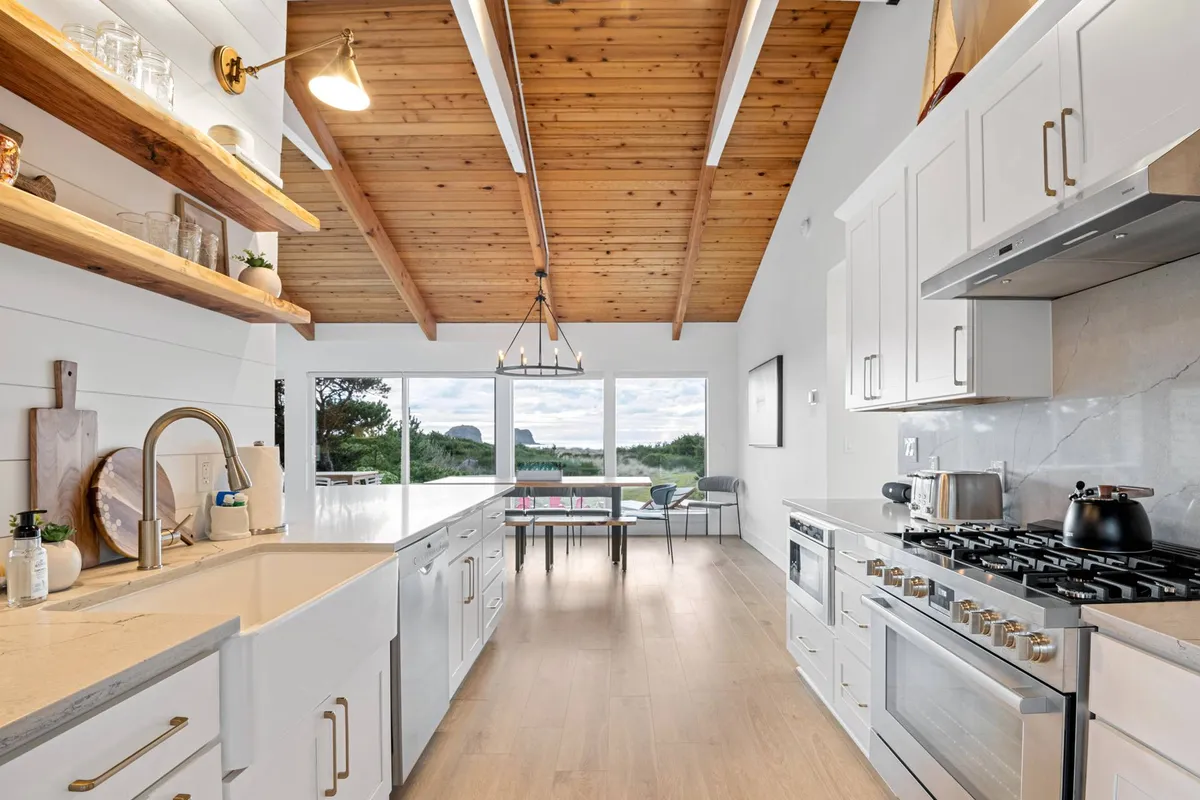 A modern kitchen with a central island, sleek appliances, exposed wooden beams, and large windows bringing in light and providing glimpses of the Pacific. The setting is ideal for preparing meals together and seamlessly connects to the home’s main living space.