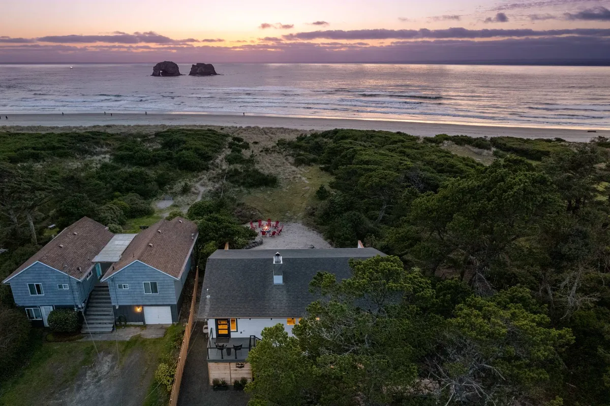 A sweeping view of the coastline with gentle waves lapping at the sandy shore, two distinctive rock formations in the water, and a fire pit area surrounded by houses. The scene feels tranquil and inviting, perfect for family gatherings and oceanfront activities.