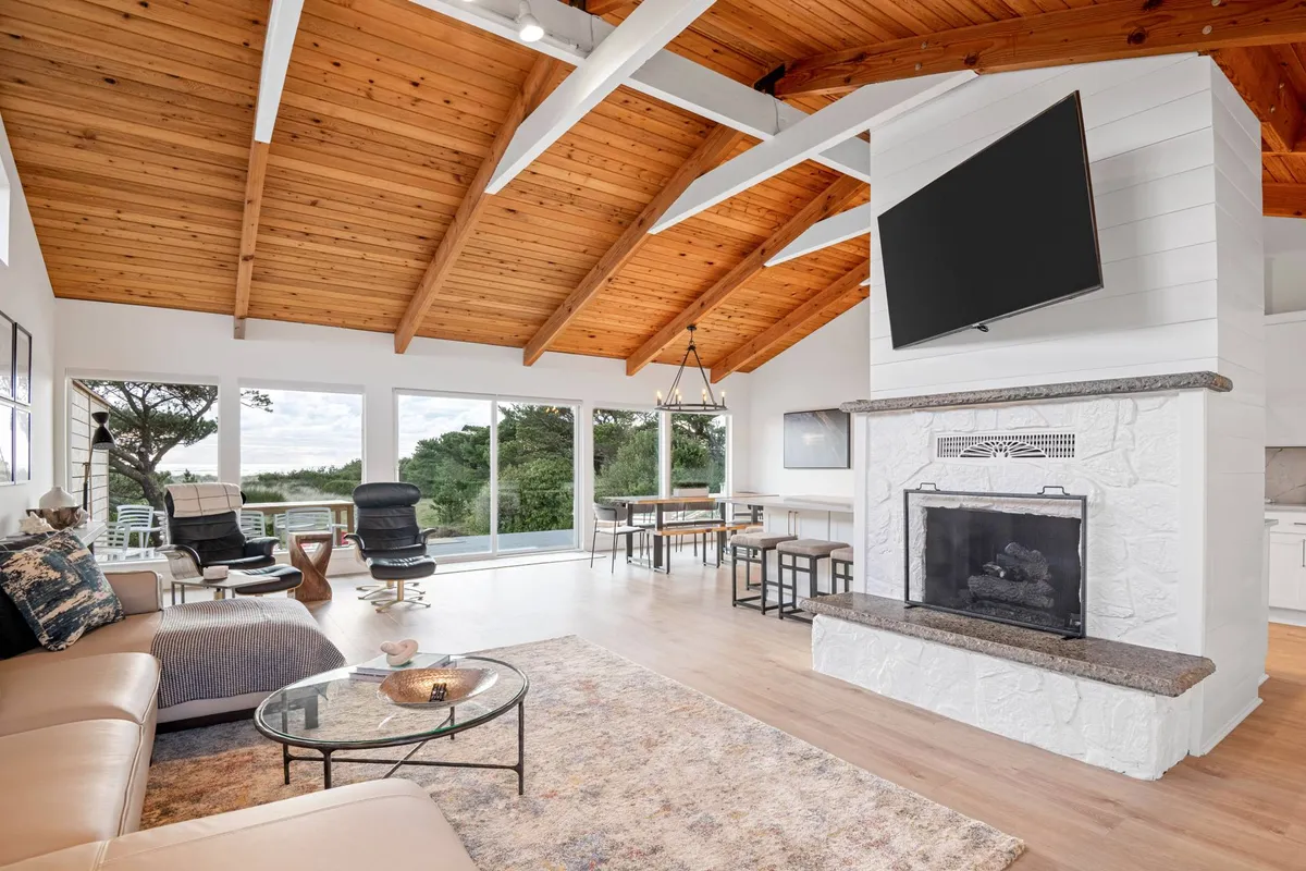 A spacious open-concept living and dining area with a prominent stone fireplace, large windows framing expansive ocean views, exposed wooden beams, and a dining table set for gatherings. Natural light floods the room, and the décor is stylish yet comfortable, encouraging relaxation or socializing.