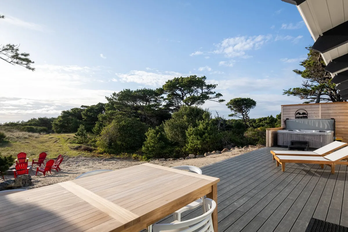 This image features a large wooden deck with red Adirondack chairs, a wooden dining table for alfresco meals, a hot tub, and surrounding greenery. The open layout showcases plenty of space for relaxing, socializing, and enjoying nature. The deck overlooks what appears to be a natural landscape, hinting at ocean views and easy access to outdoor amenities.